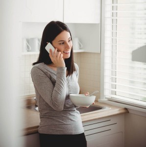 Woman talking on smart phone while holding food bowl in her kitchen with white blinds Woman talking on smart phone while holding food bowl in her kitchen with white blinds